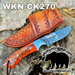 Damascus-pattern knife with an orange tooled leather sheath resting on a log; large 'WKN' on blade.