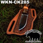 Fixed-blade knife with wooden handle in a brown embossed leather sheath on soil, text WKNC K285 at top edge.