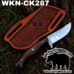 Fixed‑blade knife with a wooden handle and brown patterned leather sheath, outdoors on dirt. RB branding visible on the blade.