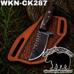 Fixed-blade hunting knife with serrated spine on a brown tooled leather sheath outdoors.