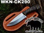 Fixed-blade knife with textured handle laid on a brown leather sheath outdoors; branding text WKN-CK290 appears above.