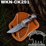 Fixed-blade knife with a marbled handle resting on an orange leather sheath outdoors by a tree, product photo for hunting gear