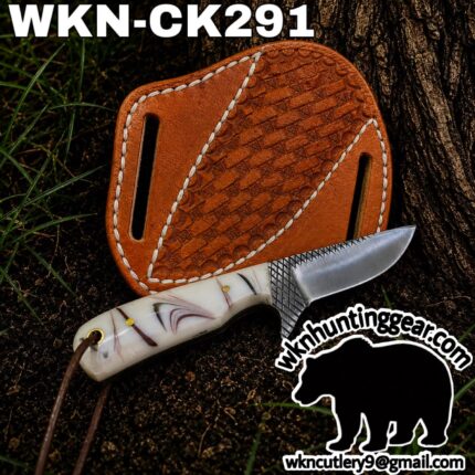Close-up of a fixed-blade knife with a patterned handle next to a tan tooled leather sheath on dirt and grass; model WKN-CK291 visible at top.