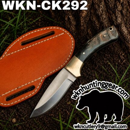 Fixed-blade knife with a dark marbled handle and brass guard, placed on dirt beside a tan leather sheath; outdoor scene with grass, featuring the wkncutlery watermark.