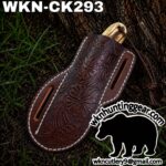Brown tooled leather knife sheath with floral pattern, shown outdoors against a tree trunk; model WKN-CK293 and huntinggear logo are visible.