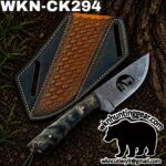 Damascus hunting knife with a textured blue/black handle beside a decorative leather sheath on dirt outdoors.