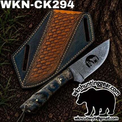 Damascus hunting knife with a textured blue/black handle beside a decorative leather sheath on dirt outdoors.