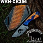 Damascus-style hunting knife with blue and wood handle beside a decorative leather sheath on grass, with a watermark/logo at the bottom right.