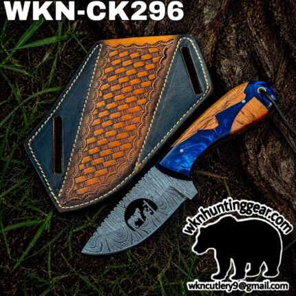 Damascus-style hunting knife with blue and wood handle beside a decorative leather sheath on grass, with a watermark/logo at the bottom right.