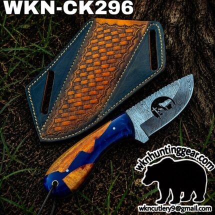 Knife with a Damascus-pattern blade and blue/orange handle beside a decorated blue leather sheath on dirt and grass.