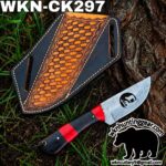 Damascus knife with red and black handle beside a brown tooled leather sheath on grass and tree trunk, outdoors gear focus.