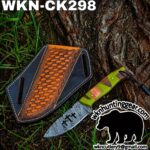 Damascus-style knife with a green-and-wood handle beside a decorative leather sheath on grass by a tree trunk.