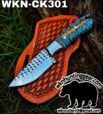 Fixed-blade knife with a blue patterned handle in a brown tooled leather sheath, laid on grass and soil.