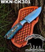 Fixed-blade hunting knife with a blue-patterned handle resting on an orange leather sheath outdoors, near a tree trunk. The image includes the site text wkn huntinggear at the bottom right.