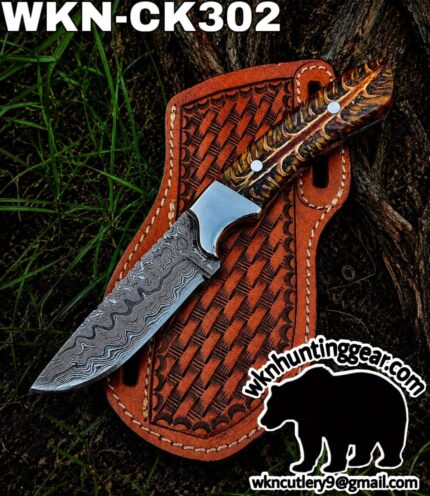 Damascus steel knife with a patterned horn handle laid on a brown tooled leather sheath outdoors; watermark with wknhuntinggear.com and a bear silhouette.