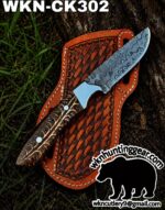 Damascus knife with textured blade resting on a brown leather sheath outdoors; branding at bottom right shows 'wknhuntinggear.com'