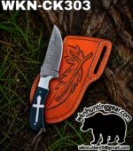 Damascus fixed‑blade knife with a dark handle and cross emblem, resting beside an orange engraved leather sheath outdoors.