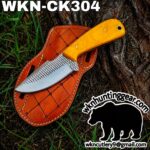 Knife with orange handle on a brown leather sheath labeled WKN-CK304 lying on tree bark outdoors; grass visible in the background as part of the scene.