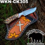 Fixed-blade knife with a carved, antler-like handle resting on a tooled leather sheath featuring sunflowers and a cross motif, outdoors on grass and tree bark.