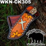 Hand-tooled leather knife sheath with orange panel, sunflower motifs and a cross, lying on grass beside a tree trunk.