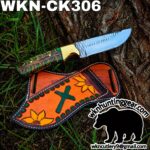 Decorative hunting knife with a green, textured handle and brass bolster lay on a tree next to a colorful leather sheath with sunflowers and a cross motif; branding for wkn hunting gear visible.