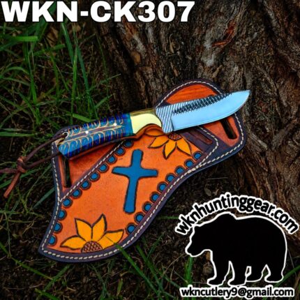 Colorful fixed-blade knife resting on an orange leather sheath decorated with sunflowers and a blue cross, placed on tree bark with grass nearby.