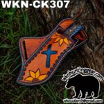 Handmade leather knife sheath in orange with blue cross and yellow sunflowers, lying on tree bark; branding and watermark show WKN Cutlery.