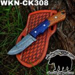 Damascus blade knife with a blue/orange patterned handle resting on a red leather sheath outdoors by a tree. Watermark shows wknhuntinggear.com and contact info.