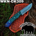 Damascus steel knife with a blue-green hexagonal handle laid across an orange tooled leather sheath outdoors on grass and tree bark.