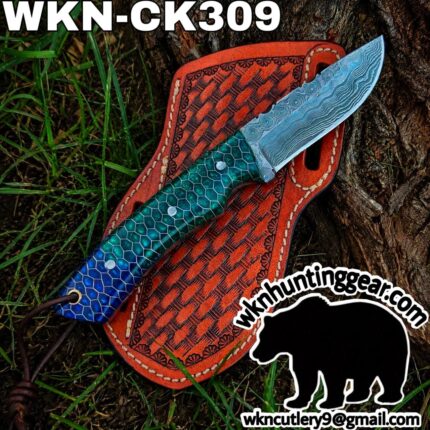 Damascus steel knife with a blue-green hexagonal handle laid across an orange tooled leather sheath outdoors on grass and tree bark.