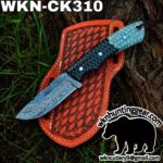 Damascus knife with a patterned blue-green handle laid on an orange leather sheath outdoors by a tree trunk.