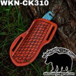Orange tooled leather knife sheath with a blue handle resting on a tree trunk; watermark logo and bear silhouette in the lower right corner