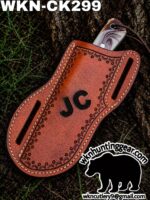 Brown tooled leather knife sheath with 'JC' initials and a visible blade, placed on grass; huntinggear logo in the corner.