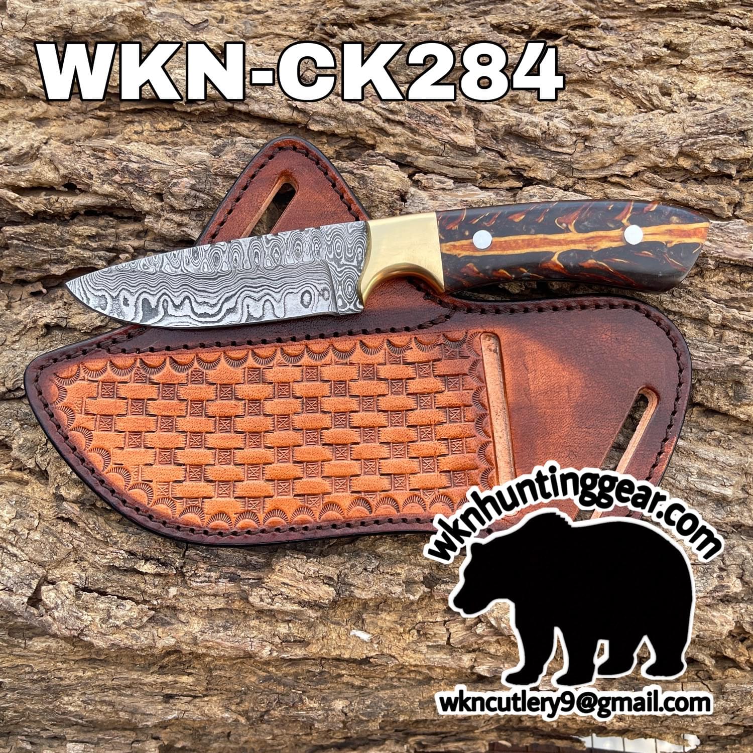 Damascus-pattern hunting knife with brass bolster and wood handle resting on a brown leather sheath against rough bark background.