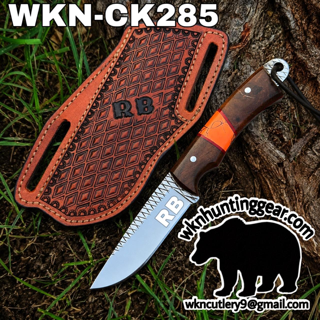 Hunting knife with a wood handle and orange wrap resting on a brown patterned leather sheath with RB branding, outdoors near grass and a tree trunk.