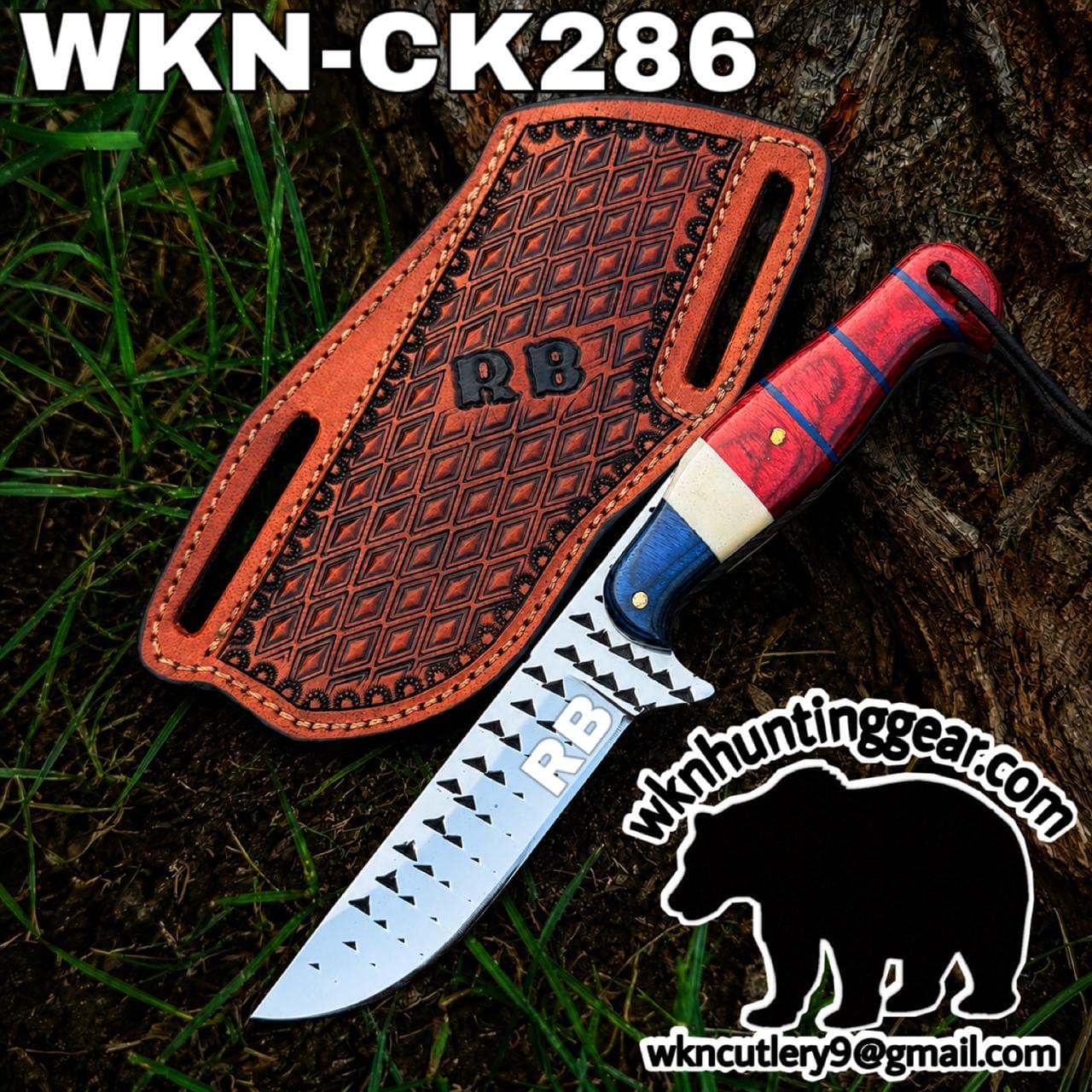 Hunting knife with a patterned blade and red handle, resting beside a brown leather sheath on grass (branding visible).