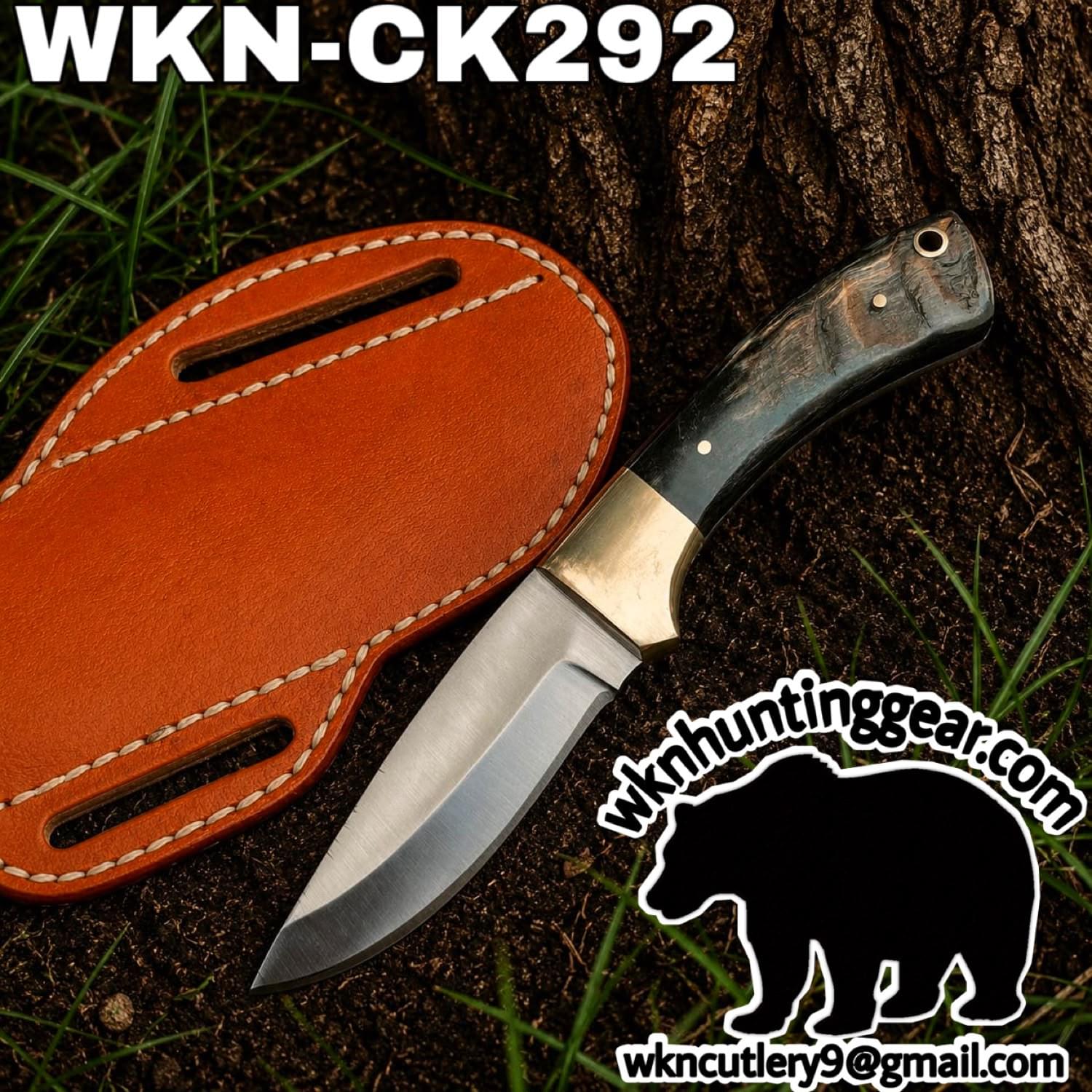 Fixed-blade knife with a dark marbled handle and brass guard, placed on dirt beside a tan leather sheath; outdoor scene with grass, featuring the wkncutlery watermark.