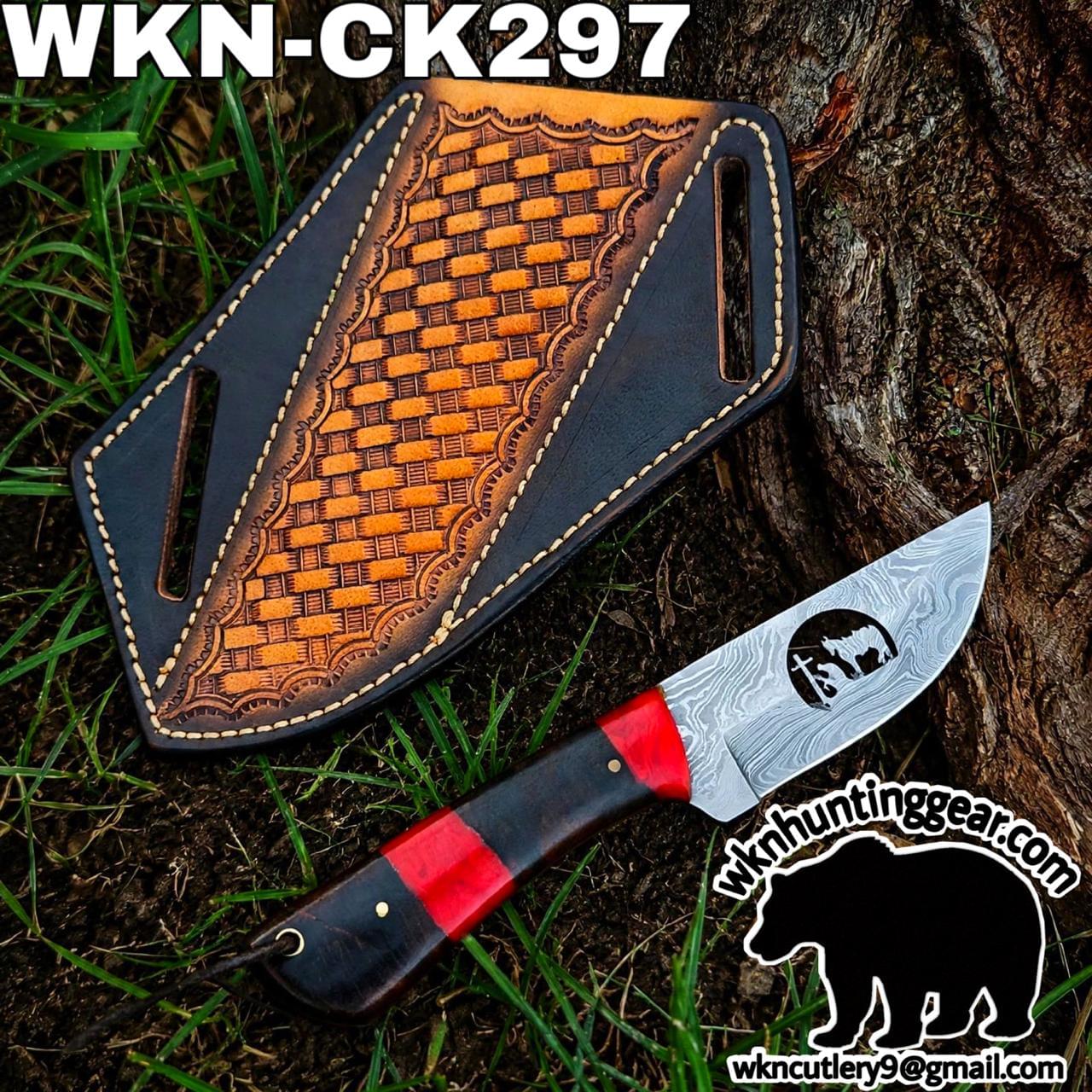 Damascus knife with red and black handle beside a brown tooled leather sheath on grass and tree trunk, outdoors gear focus.