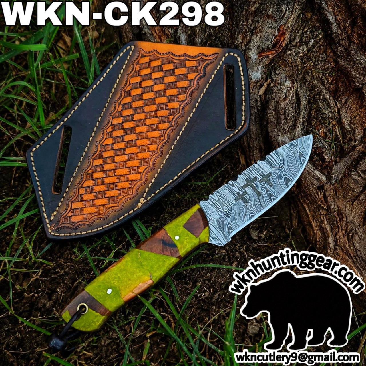 Damascus knife with a green and brown handle lying on grass beside a brown leather sheath by a tree; watermark with a bear logo and email bottom-right.