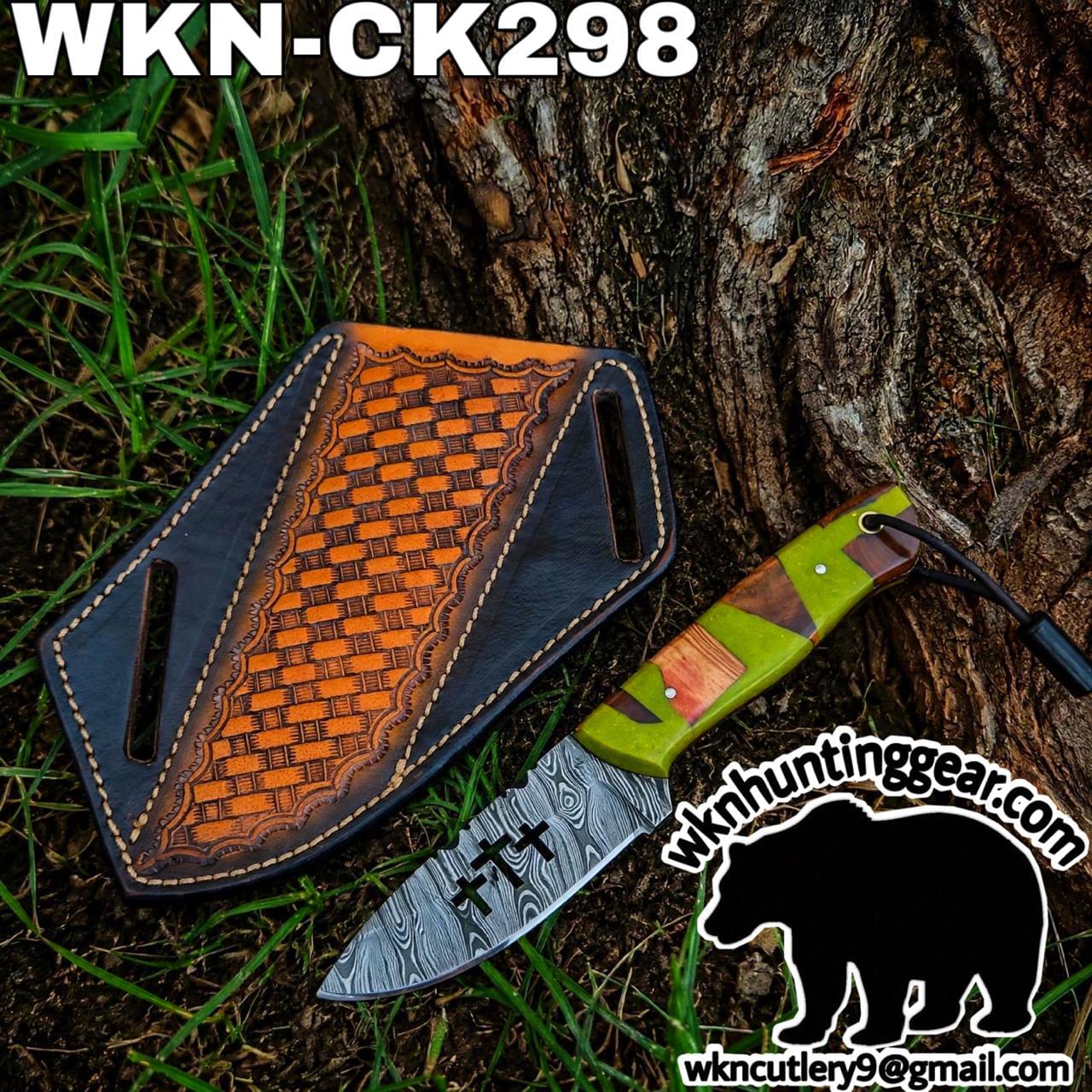 Damascus-style knife with a green-and-wood handle beside a decorative leather sheath on grass by a tree trunk.