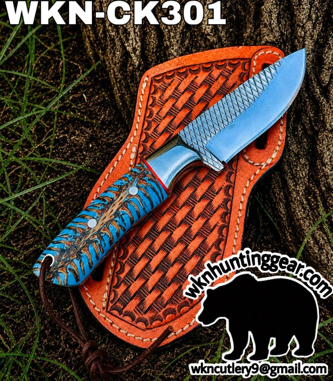 Fixed-blade hunting knife with a blue-patterned handle resting on an orange leather sheath outdoors, near a tree trunk. The image includes the site text wkn huntinggear at the bottom right.