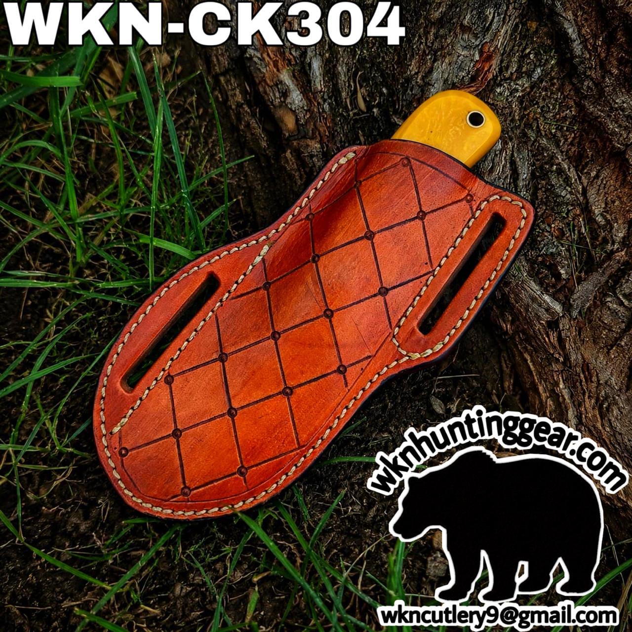 Orange-brown leather knife sheath with white stitching lying on grass beside a tree trunk, yellow knife handle peeking from the top; watermark for wkn hunting gear visible in corner.