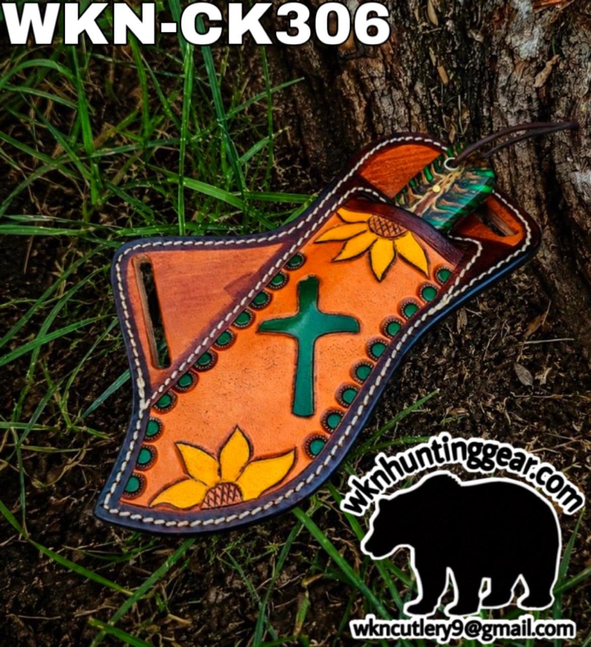 Decorative orange leather knife sheath with a green cross and sunflower motifs, white stitching, on grass next to a tree; watermark with bear logo and email.