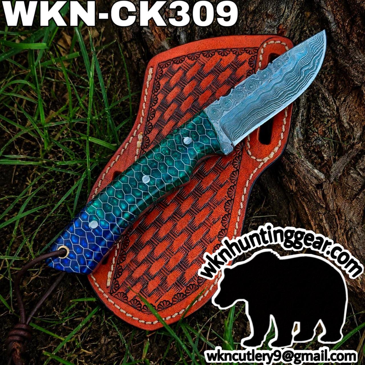 Damascus steel knife with a blue-green hexagonal handle laid across an orange tooled leather sheath outdoors on grass and tree bark.