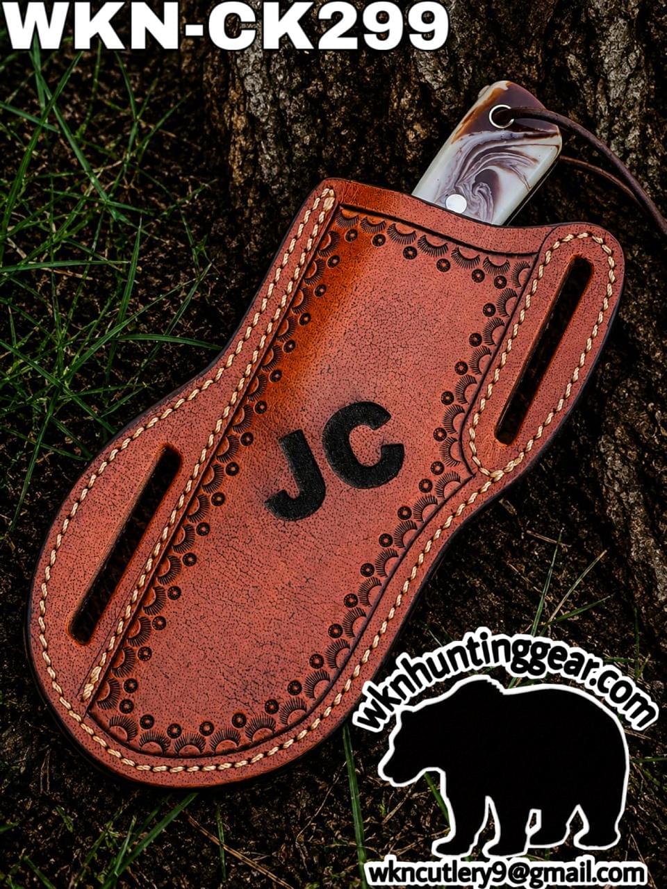 Brown tooled leather knife sheath with 'JC' initials and a visible blade, placed on grass; huntinggear logo in the corner.